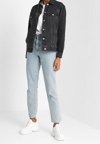 Person som bär en svart jeansjacka, vit t-shirt, ljusblå högmidjade jeans och vita klumpiga sneakers, stående mot en enfärgad bakgrund.