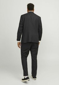 Costume noir à coupe ajustée, avec un pantalon slim et une veste légèrement plus longue. Porté avec des baskets noires et des chaussettes blanches.