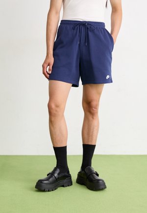 Osoba nosząca granatowe spodenki Nike, czarne skarpetki i czarne masywne buty stoi na zielonej podłodze na białym tle.