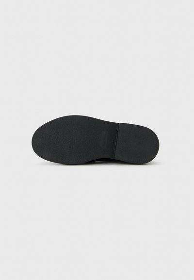 Suola di scarpa nera con texture liscia e leggero tacco, vista dal basso su uno sfondo chiaro e uniforme.