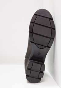 Semelle de chaussure noire avec motif de bande de roulement texturé et segmenté, taille 37, positionnée à la verticale contre un fond blanc en angle.