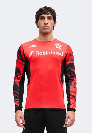 Homme portant un maillot de sport rouge et noir à manches longues avec le logo Robinhood et le blason de l'équipe, debout devant un fond uni.