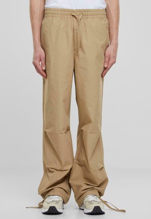 Beige, loose-fitting pants with an elastic waistband and drawstring. Fabric appears lightweight with a smooth texture. Ankle cuffs are adjustable.