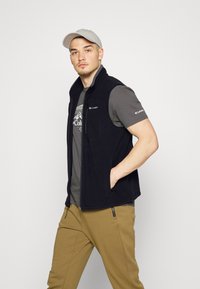 Schwarze Fleece-Weste mit Reißverschluss, ausgestattet mit zwei seitlichen Taschen, über einem grauen T-Shirt getragen, kombiniert mit khakifarbenen Hosen und einer hellgrauen Mütze.
