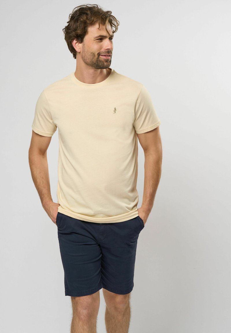 Tricou din bumbac crem cu guler rotund și mâneci scurte, având un mic logo brodat pe piept; asortat cu pantaloni scurți bleumarin.
