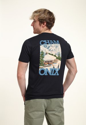 Muž v černém tričku s motivem horské chaty zasypané sněhem a nápisem „CHAM ONIX“ na zádech.