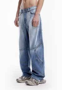 Svijetloplave traperice širokog kroja, s detaljima zakrivljenih šavova i pojasom srednjeg struka. Nosi sivi tenisice.