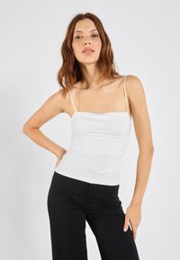 White fitted camisole top with thin straps, featuring a smooth texture and a square neckline, paired with black wide-leg pants.