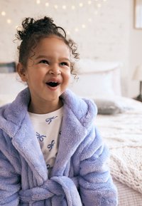 Peignoir en peluche violet clair avec un col châle, ceinturé à la taille, porté par-dessus une chemise blanche ornée d'un motif de nœuds bleus.