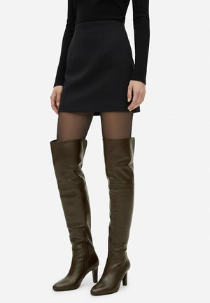 Mujer con minifalda negra, medias negras transparentes y botas de cuero oscuras hasta la rodilla con tacón.