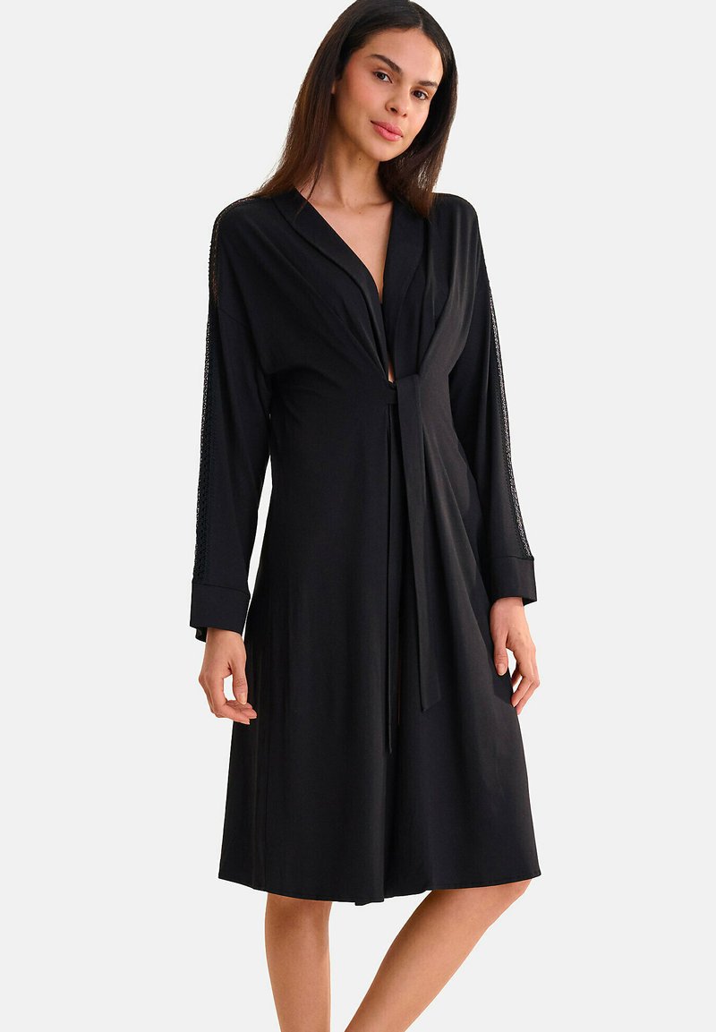 Robe noire cintrée avec manches longues et fermeture à nouer. Elle possède une texture lisse et des coutures subtiles le long des bords des manches.