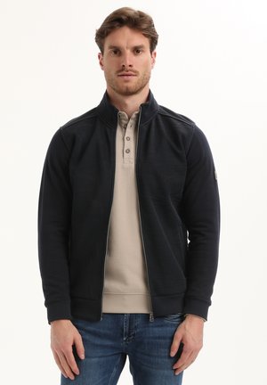 Man met lichtbruin haar, gekleed in een zwarte ritsjack over een beige knoopshirt en blauwe spijkerbroek, staande tegen een witte achtergrond.