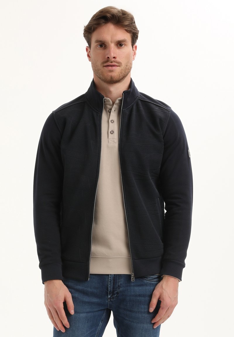 Hombre con cabello castaño claro que lleva una chaqueta negra con cremallera sobre una camisa beige abotonada y jeans azules, de pie contra un fondo blanco.