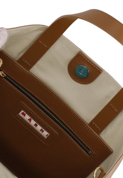 Borsa in pelle marrone con interno in tessuto canvas, tasca con zip e chiusura a scatto. Presenta un piccolo bottone teal e una toppa rossa con il logo.