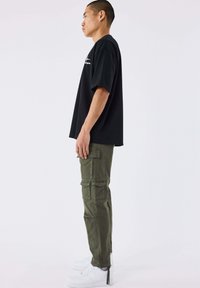 Man staat zijwaarts, draagt een zwart oversized T-shirt, olijfgroene cargobroek met zakken, witte sneakers en een zilveren ring aan de linkerhand.