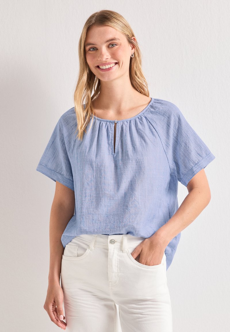Femme souriante portant un chemisier ample bleu clair à manches courtes et un pantalon blanc, debout avec une main dans la poche devant un fond uni.