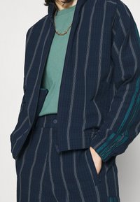 Marinblå randig jacka med matchande byxor, i en strukturvävd tyg med teal accenter, tillsammans med en grön skjorta och ett guldhalsband.