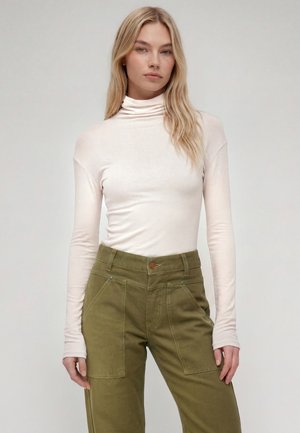Femme aux longs cheveux blonds, portant un col roulé blanc et un pantalon taille haute vert olive, debout devant un fond uni.