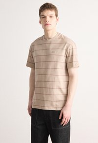 Beige puuvillane T-särk, millel on horisontaalsed helepruunid triibud, ümmargune kaelus, lühikesed varrukad ja väike logo patch rinna lähedal.