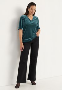 Teal sammetslik topp med V-ringning, avslappnad passform och korta ärmar, kombinerad med svarta utsvängda byxor och svarta öppna skor.