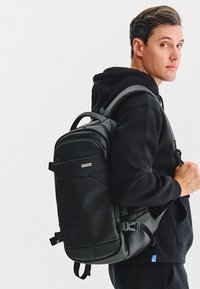 Czarny plecak wykonany z gładkiego materiału, z regulowanymi paskami, zamykaną na zamek kieszenią z przodu oraz subtelnym logo po boku.