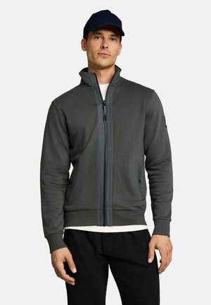 Mann trägt eine dunkelgraue Jacke mit Reißverschluss über einem weißen Hemd, schwarze Hose und eine dunkelblaue Baseballkappe, steht vor einem hellen Hintergrund.