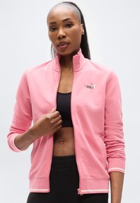Ελαφρώς ροζ ζακέτα με φερμουάρ και μακριά μανίκια, με ψηλό γιακά, ριμπ μανσέτες και λογότυπο Puma στο στήθος. Μαύρο αθλητικό τοπ από κάτω.