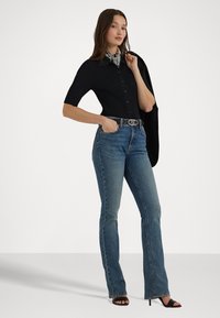 Camisa negra de botonadura con ribetes y mangas cortas, jeans acampanados azules y tacones negros con tiras. Destaca un cinturón plateado y un detalle de bufanda en el cuello.