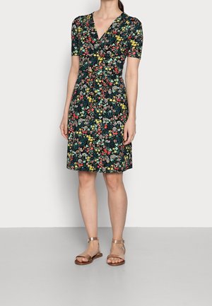 Kvinde iført en knælængde, kortærmet blomsterkjole med V-hals og beige flade sandaler med ankelrem, står foran en ensfarvet baggrund.
