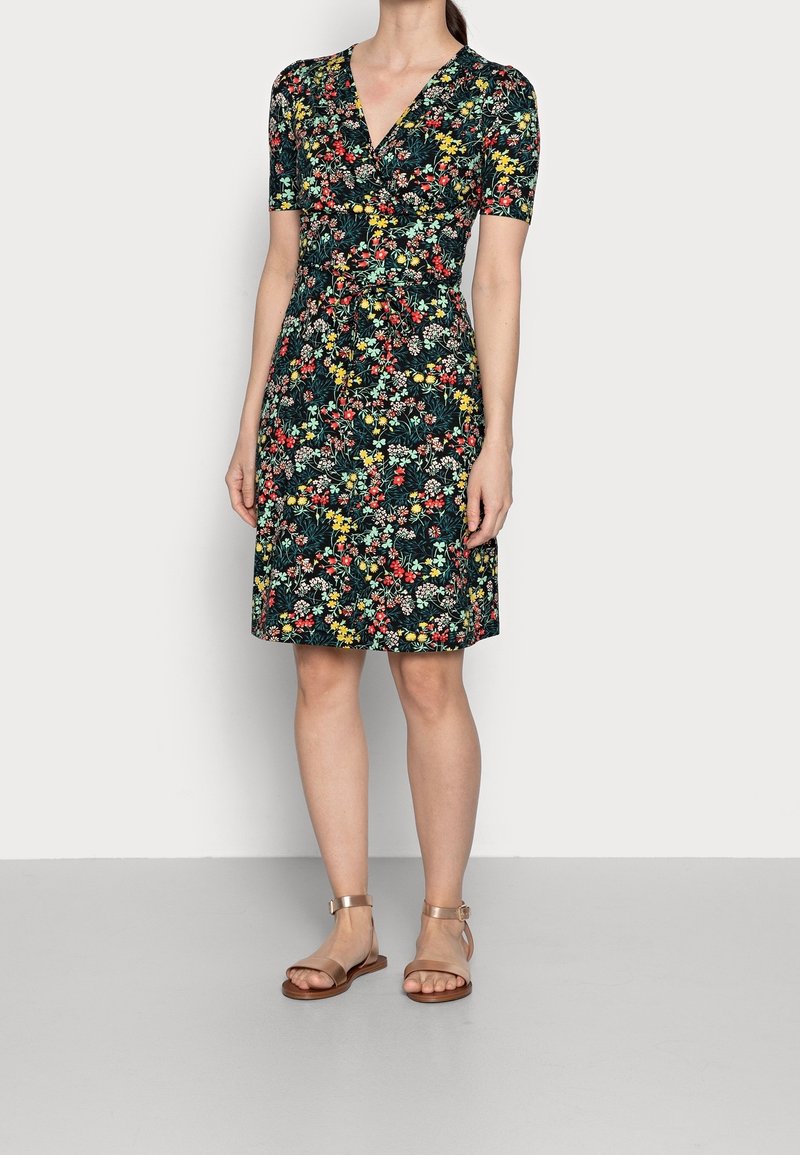 Femme portant une robe à fleurs courte aux genoux, manches courtes, col en V, et des sandales plates beiges avec bride à la cheville, debout devant un fond uni.
