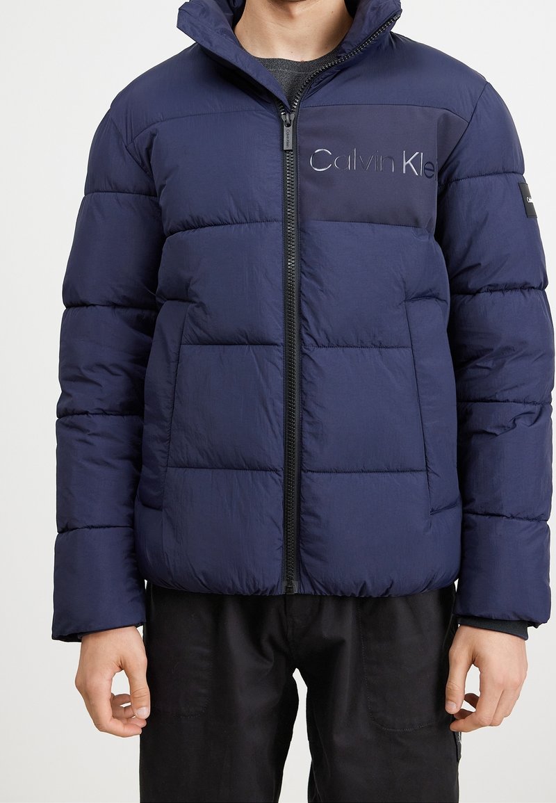 Mörkblå dunjacka med dragkedja, prydd med en logotyp på bröstet, två framfickor och hög krage. Slät textur och quiltad design.