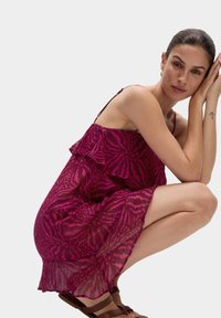 Abito fucsia trasparente con volant sovrapposti, caratterizzato da una fantasia a zebra. Spalline sottili e orlo a campana valorizzano il design. Indossato con sandali marroni.