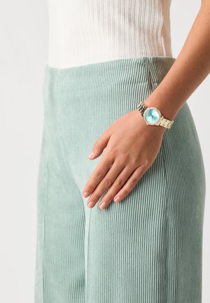 Gold-tone watch with a light blue face, adorned with rhinestones, displayed on a hand near light green ribbed corduroy pants.