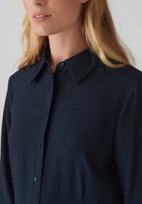 Camicia blu navy con colletto, caratterizzata da una texture liscia e maniche lunghe. Bottoni neri completano il design minimalista.