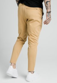 Pantaloni slim-fit color sabbia con una texture liscia, dotati di una tasca posteriore e indossati con sneaker bianche. Il design enfatizza una silhouette moderna.