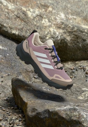 Zapatilla de trail running con diseño en beige, rosa y blanco, suela negra y cordones, situada sobre terreno rocoso al aire libre.