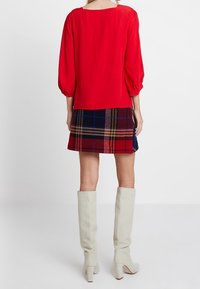 Rode blouse met pofmouwen, gecombineerd met een geruite mini-jurk in rood, blauw en geel. Witte kniehoge haklaarzen maken de look compleet.