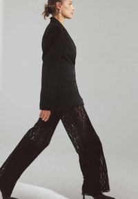 Svart skräddarsydd kavaj med en enda knapp, kombinerad med vida, genomskinliga byxor i spets som har intrikata mönster och en mjuk textur.