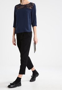 Blusa blu navy con pannello di pizzo sullo scollo, maniche a ¾; abbinata a pantaloni neri a vita alta e mocassini neri lucidi.