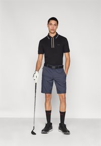 Črna polo majica z belim in bež ovratnikom, mornarsko modre kratke hlače, črni čevlji, črno-modre črtaste nogavice, drži golf palico in roko z rokavico.