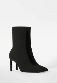 Botas negras de media pantorrilla, con punta afilada y tacones de aguja, con parte superior de tela suave, exhibidas una al lado de la otra sobre un fondo blanco liso.