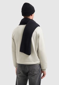 Marineblauwe ribgebreide beanie en sjaal over een lichtgrijze sweatshirt gedrapeerd, gedragen met donkergrijze jeans, gezien vanuit de achterkant.