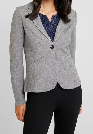 Kvinde iført en tætsiddende grå blazer over en navy mønstret top og sorte bukser, stående mod en lysegrå baggrund.
