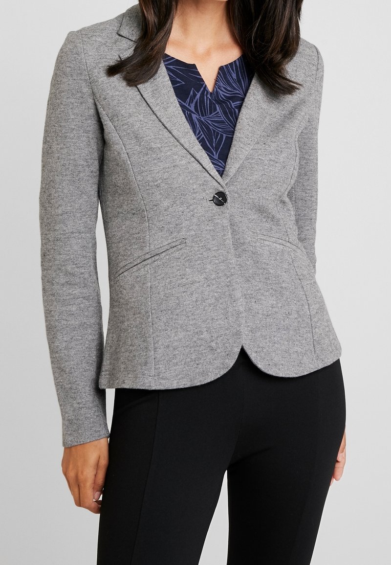 Femme portant un blazer gris ajusté sur un haut bleu marine à motifs et un pantalon noir, debout sur un fond gris clair.