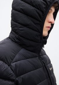 Veste matelassée noire avec un design matelassé, un col montant et une capuche. Fabriquée en tissu léger et texturé avec des détails de fermeture éclair.