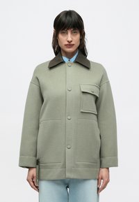 Zeleni sukljasti suknjič z mešanico volne z rjavim ovratnikom, sprednjimi žepi in zapiranjem na gumbe. Ima sproščen kroj in subtilno teksturo, kombiniran s svetlomodro srajco spodaj.