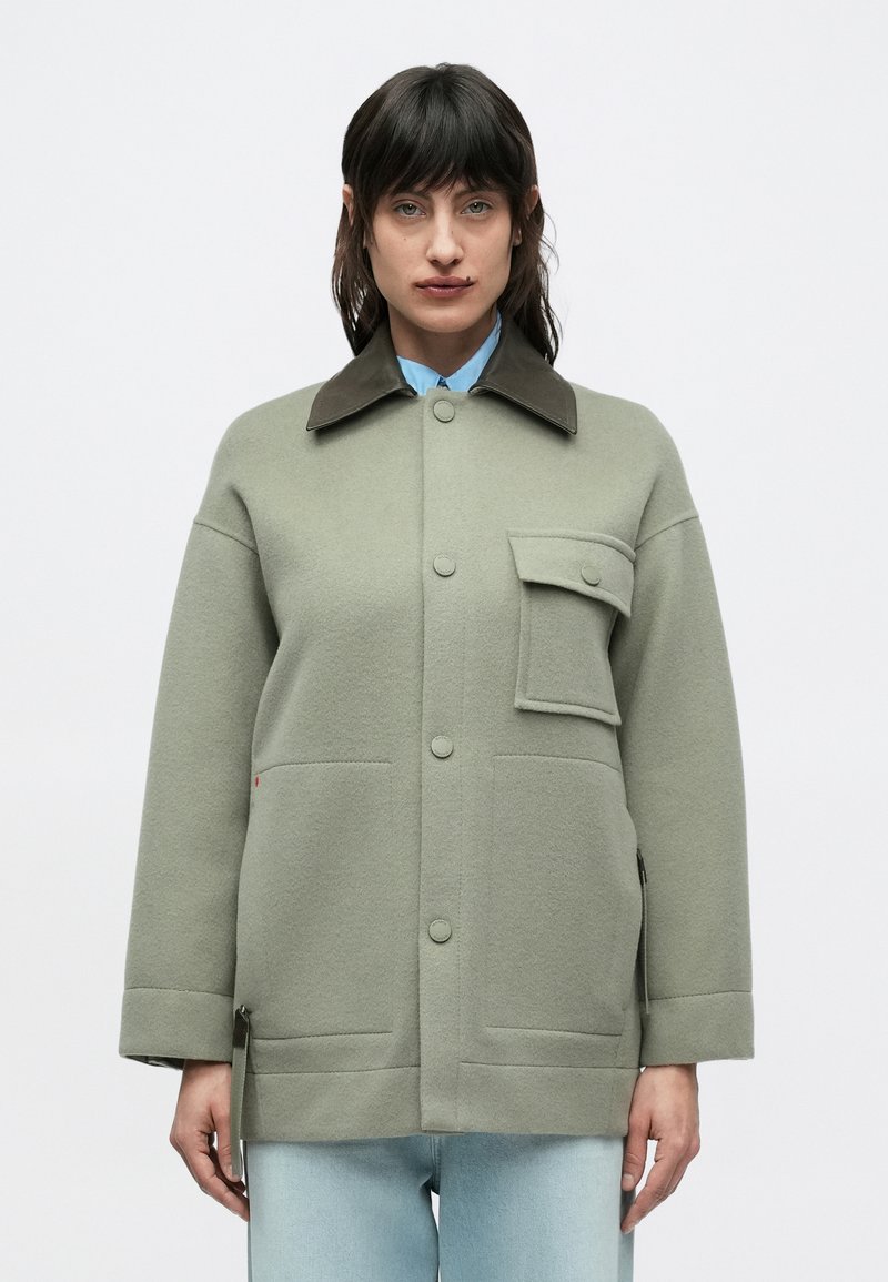 Zeleni sukljasti suknjič z mešanico volne z rjavim ovratnikom, sprednjimi žepi in zapiranjem na gumbe. Ima sproščen kroj in subtilno teksturo, kombiniran s svetlomodro srajco spodaj.