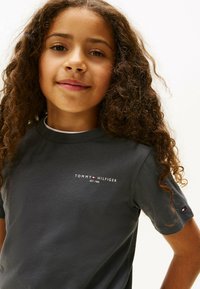 Pige med krøllet hår iført en mørkegrå Tommy Hilfiger t-shirt med logo på brystet og ærmet, som smiler svagt mod en hvid baggrund.