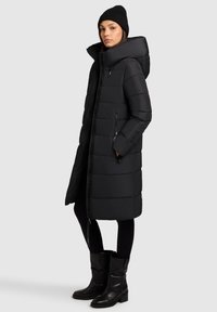 Čierny zateplený kabát s vysokým golierom, prešívaným dizajnom a zipsovými vreckami. Nosí sa s čiernymi legínami a čižmami po kolená.