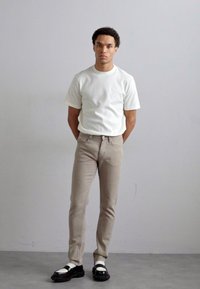 Hvid ribbet kortærmet t-shirt, beige slim-fit bukser og sorte sandaler med hvide remme. Enkel, moderne design mod en ensfarvet baggrund.